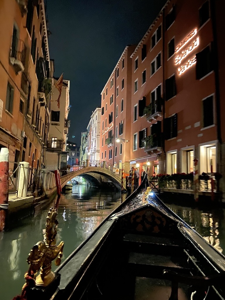 A bord d'une gondole de nuit sur les canaux de Venise. Les lumières des immeubles se reflètent sur l'eau.